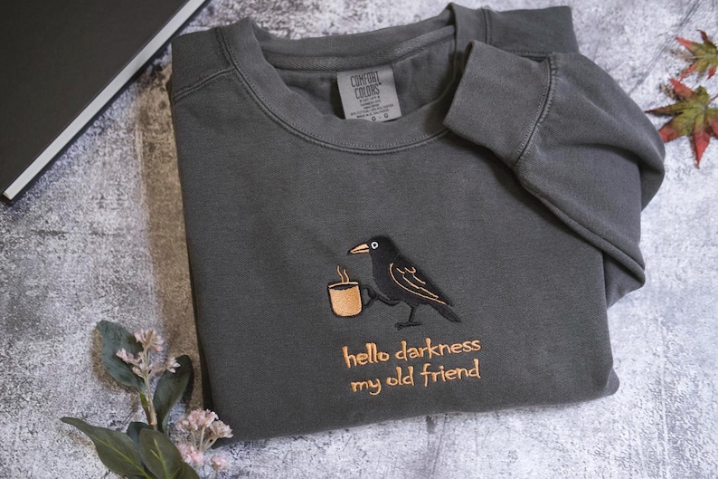 a gray sweatshirt with a black bird embroidered on it, holding a cup, and the text "Hello darkness, my old friend" printed on it.