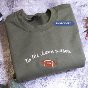Könnte beinhalten: Olivgrünes Sweatshirt mit dem gestickten Schriftzug "tis the damn season" und einer Football-Grafik. Das Wort "EMBROIDERY" steht in einem blauen Banner. Das Sweatshirt liegt gefaltet auf einer strukturierten Oberfläche.