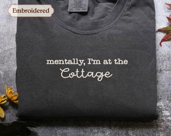 May include: A dark gray, folded t-shirt with the embroidered text "mentally, I'm at the Cottage" in white script. The shirt has a crew neck and a tag that reads "Comfort Colors". The shirt is labeled "Pepper - Comfort Colors Shirt".