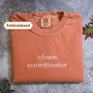 May include: A terracotta-coloured crew neck t-shirt with the embroidered text "chaos coordinator" in white thread on the chest. The shirt features a Comfort Colors brand label at the inner neckline, which indicates a size medium and 100% cotton material.