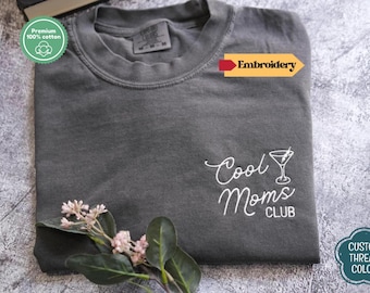 May include: A dark gray t-shirt with the embroidered text "Cool Moms Club" and a cocktail glass design. The shirt is made of premium 100% cotton. A sprig of flowers is placed near the bottom of the shirt.