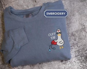 May include: A blue-grey sweatshirt with an embroidered design of a cartoon seagull wearing sunglasses and a nurse's hat, holding a medical bag and stethoscope. The text "CUFF 'EM" is above the seagull. The word "EMBROIDERY" is in a blue banner.