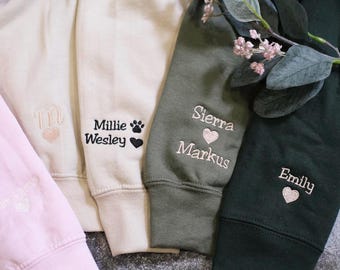 May include: Four different colored sweatshirts with embroidered names and designs. The sweatshirts are pink, beige, green, and black. The names on the sweatshirts are Millie Wesley, Sierra Markus, Emily, and a monogram.
