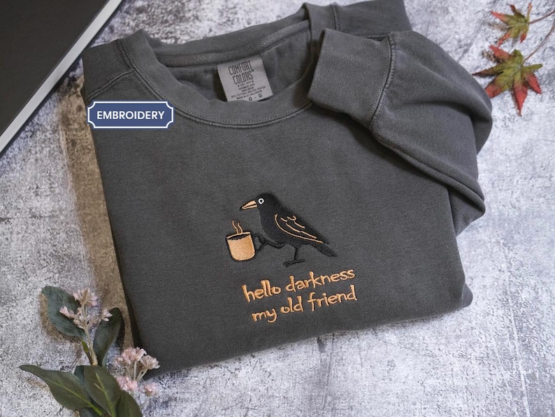 a gray sweatshirt with a black bird embroidered on it, along with the text "Hello darkness, my old friend" printed on it. The sweatshirt is placed on a gray surface, and there are some leaves and a book visible in the background.