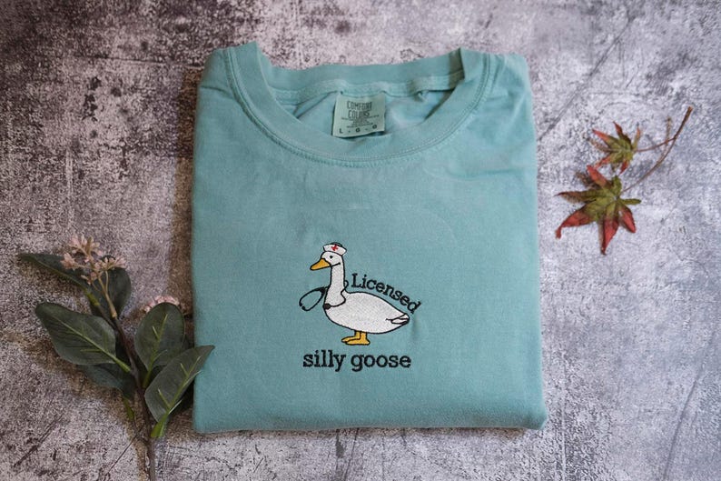 A light blue t-shirt with a cartoon image of a duck wearing a nurse's outfit and the text "Licensed silly goose" printed on it, along with some leaves and a small flower.