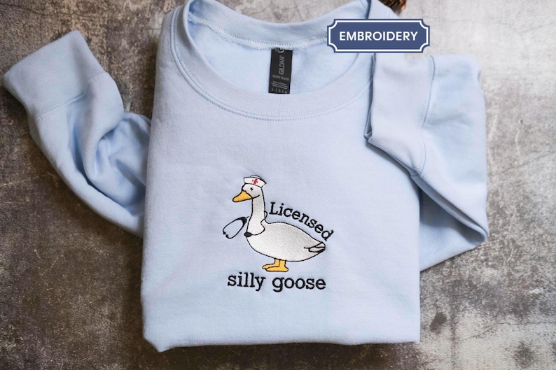 A light blue t-shirt with a cartoon image of a duck wearing a nurse's cap and the text "Licenced silly goose" printed on it.