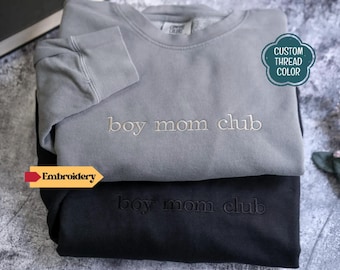 May include: A grey crewneck sweatshirt and a black crewneck sweatshirt, both featuring the embroidered text "boy mom club" on the chest. The grey sweatshirt has light-coloured embroidery, while the black sweatshirt has matching black embroidery.