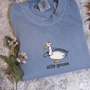 A blue t-shirt with a cartoon image of a white goose wearing a nurse's cap and the text "Licensed silly goose" printed on it, along with some flowers and a leaf.