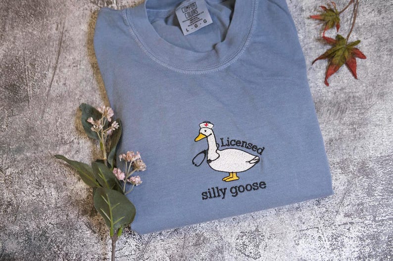 A blue t-shirt with a cartoon image of a white goose wearing a nurse's cap and the text "Licensed silly goose" printed on it, along with some flowers and a leaf.