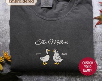 May include: A charcoal gray crewneck sweatshirt featuring white embroidery. The text reads "The Millers" above two white ducks, one wearing a black top hat and bow tie, the other a white veil. The text "EST" and "2026" flank the ducks.