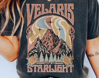 May include: A dark gray t-shirt with a vintage graphic design featuring a mountain range, sunflowers, and the text "Velaris city of Starlight".