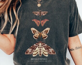 Puede incluir: Camiseta gris oscuro con un gráfico de varias mariposas y polillas en varios colores. El texto "AMATEUR LEPIDOPTERIST" y "THE STUDY OF BUTTERFLIES AND MOTHS" están impresos debajo. La camiseta se combina con vaqueros azules y zapatillas blancas.