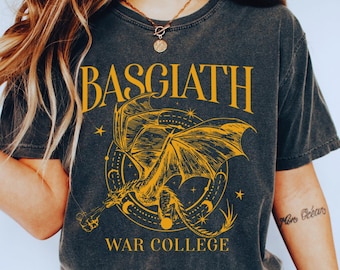 May include: Dark gray t-shirt with the text "BASGIATH WAR COLLEGE" in gold lettering. A gold dragon graphic is centered below the text. The shirt is worn with blue jeans and white shoes.