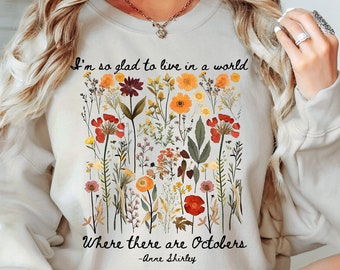 May include: Cream-colored sweatshirt featuring a floral design with red, orange, and yellow flowers. The text reads, "I'm so glad to live in a world where there are Octobers" by Anne Shirley. The sweatshirt is a comfortable, casual style.