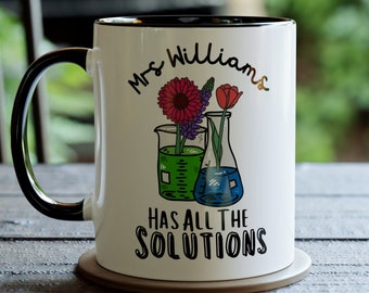 Puede incluir: Taza de cerámica blanca con asa y borde negros, con el texto "Mrs. Williams HAS ALL THE SOLUTIONS". La taza tiene una colorida ilustración de flores en vasos de precipitados y matraces. La taza está sobre una mesa de madera.