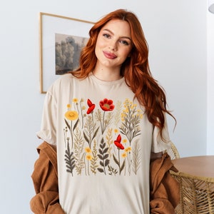 May include: A beige t-shirt with a floral print featuring red and yellow flowers. The flowers are surrounded by green leaves and stems.