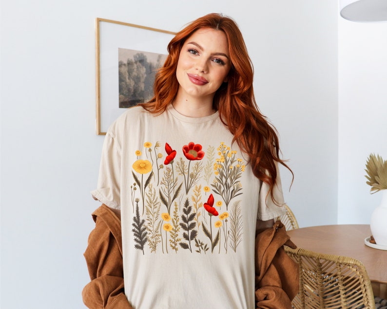 May include: A beige t-shirt with a floral print featuring red and yellow flowers. The flowers are surrounded by green leaves and stems.