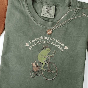 Peut inclure: T-shirt vert olive avec un dessin d'une grenouille à vélo, portant un chapeau et tenant des fleurs. Le texte "Embarking on some good old Irish mischief" est imprimé sur le t-shirt. Un collier doré est posé sur le t-shirt.
