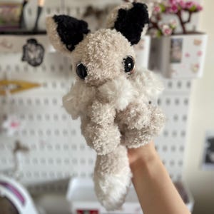 May include: A handmade plush toy fox with large, black-rimmed eyes and black-tipped ears. The fox is primarily beige with white accents on its chest and tail. The soft, fluffy texture gives it a cuddly appearance.