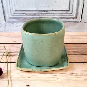 Peut inclure: Un ensemble tasse et soucoupe en céramique vert clair. La tasse est cylindrique avec un bord légèrement évasé, et la soucoupe est carrée avec des coins arrondis. L'ensemble est posé sur une surface en bois.