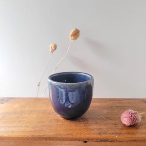 Peut inclure: Une petite tasse en céramique arrondie avec un vernis bleu indigo profond et brillant. Le récipient présente une silhouette lisse et incurvée avec une base légèrement effilée et un bord large et ouvert, révélant une surface réfléchissante.