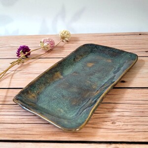 Peut inclure: Plateau de service rectangulaire en céramique avec un glaçage vert d'eau et bronze. Le plateau a des bords légèrement incurvés et une finition brillante. Fond en bois clair avec des fleurs séchées.