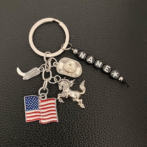 Könnte beinhalten: Silberfarbener Schlüsselanhänger mit Charms zum Thema Cowboy. Enthält einen Cowboy-Stiefel, einen Cowboyhut, ein Pferd und eine amerikanische Flagge. Der Schlüsselanhänger hat auch eine Perlenkette mit der Aufschrift "NAME".