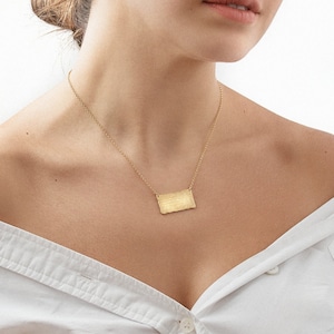 Puede incluir: Una mujer con una camisa blanca de hombros descubiertos con un collar de oro y un pequeño colgante de oro. El colgante es de forma rectangular con una superficie texturizada.