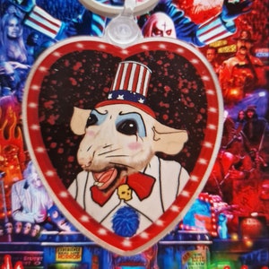 Può includere: Un portachiavi con un ciondolo a forma di cuore. Presenta un ratto dei cartoni animati vestito da Zio Sam, su uno sfondo a tema horror. Il testo "House of 1000 Corpses" è visibile.