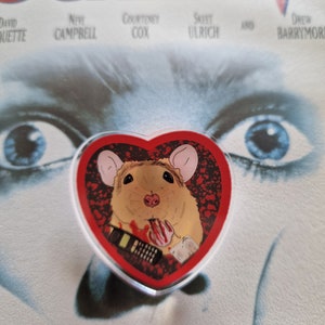 Könnte beinhalten: Eine rote herzförmige Anstecknadel mit weißem Rand. Die Anstecknadel zeigt ein Cartoon-Bild einer Ratte, die ein blutiges Telefon hält. Die Ratte trägt ein weißes Stirnband. Der Hintergrund der Anstecknadel ist rot mit schwarzen Herzen.