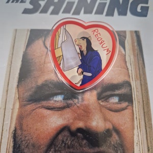 Puede incluir: Un pin en forma de corazón con un personaje de dibujos animados de zarigüeya con un kimono azul sosteniendo una cuchilla, con la palabra "REDRUM" escrita en él. El pin está sobre un fondo del cartel de la película "The Shining".