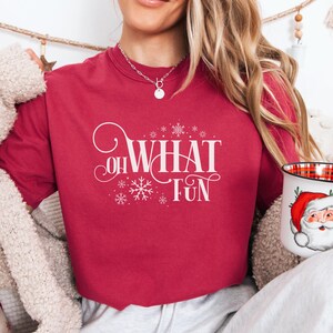 May include: A maroon t-shirt with the words "OH WHAT FUN" in white script and snowflake accents. A white mug with a Santa Claus design and a red and black plaid interior is also visible.