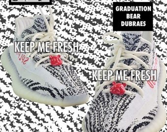 May include: Pair of white and black patterned trainers with white laces and a red bear charm. The text "KEEP ME FRESH" is printed on the side. The text "GRADUATION BEAR DUBRAES" is also visible. The shoes are on a black and white background.