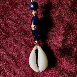 May include: Copper-colored hair accessory featuring a coiled top, a silver star charm, and a series of beads in blue and gold. A white cowrie shell hangs at the bottom. The accessory is designed for hair styling.