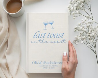 Könnte beinhalten: Ein cremefarbenes Buch mit dem Text "last toast on the coast" in Hellblau sowie zwei Cocktailgläsern. Das Buch trägt die Aufschrift "Olivia's Bachelorette" mit Datum und Ort. Eine weiße Kaffeetasse und weiße Blumen sind ebenfalls im Bild.