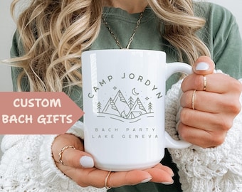 May include: White ceramic mug with the text "CAMP JORDYN" above a mountain and tent design. Below the design are the words "BACH PARTY LAKE GENEVA". The mug is held in front of a person wearing a green shirt and a white sweater. The text "CUSTOM BACH GIFTS" is on the left.