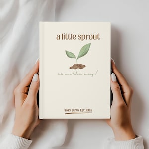 May include: A cream-colored journal held by hands, featuring the text "a little sprout is on the way!" in brown and green. A watercolor illustration of a sprout is above the text. The bottom of the journal reads "BABY SMITH EST. 2026".