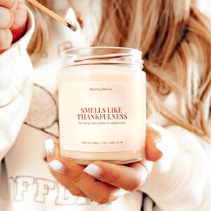 May include: A lit candle in a clear glass jar held by a person. The candle has a cream-colored wax and a label that reads "SMELLS LIKE THANKFULNESS." The text "You bring hope where it's needed most" is also on the label. The candle is being lit with a match.