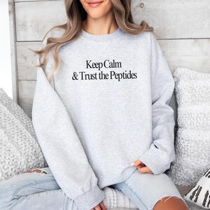 Puede incluir: Sudadera gris claro con el texto negro "Keep Calm & Trust the Peptides". La sudadera de manga larga tiene cuello redondo. La modelo lleva vaqueros azules.
