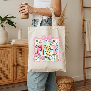 May include: Cream-colored tote bag with a colorful Pre-K design. The design includes a pink bow, daisies, stars, a ruler, and a pencil. The text "pre-k" is in a playful font with yellow, pink, and blue colors.