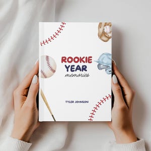 May include: A white baseball-themed memory book held by a person. The cover features a baseball, glove, helmet, and bat illustrations, with the text "ROOKIE YEAR memories" in red and blue. The name "Tyler Johnson" is also printed on the cover.