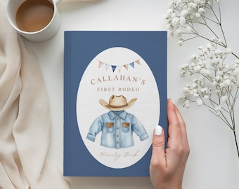 Puede incluir: Un libro de recuerdos azul con temática de vaquero. La portada presenta un diseño ovalado con una ilustración de sombrero y camisa de vaquero, el texto "Callahan's First Rodeo" y las palabras "Memory Book". Una taza de café blanca está en el fondo.