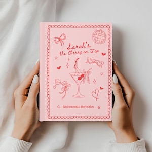 Op de afbeelding: Een roze notitieboek met een rode rand en illustraties. De cover leest "Sarah's the Cherry on Top" met een martiniglas, harten en andere afbeeldingen. De tekst "Bachelorette Memories" is ook opgenomen.