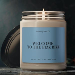 May include: A beige soy candle with a blue label that reads "Blooming Bees Co. WELCOME TO THE FIZZ BIZZ 100% SOY CANDLE | 9OZ | MADE IN USA".
