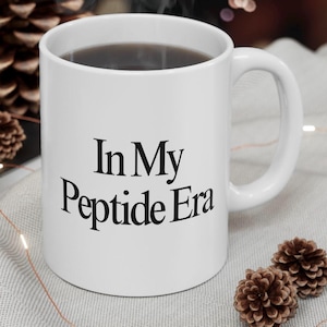 Puede incluir: Taza de cerámica blanca con el texto "In My Peptide Era" en negro. La taza está llena de un líquido oscuro y tiene un asa curvada. El fondo incluye piñas y un entorno borroso de tonos neutros.