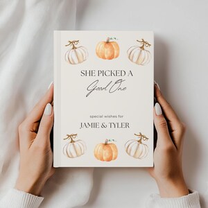 May include: A cream-coloured guest book featuring watercolour pumpkins. The cover reads "SHE PICKED A Good One" and "special wishes for JAMIE & TYLER". The book is held open, displaying the cover design.