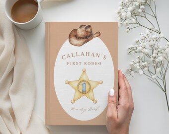Puede incluir: Un libro de recuerdos de color beige con el texto "Callahan's First Rodeo" y una ilustración de estrella de sheriff. La portada también presenta una ilustración de sombrero de vaquero. El libro está sostenido por una mano, con una taza de café y flores en el fondo.