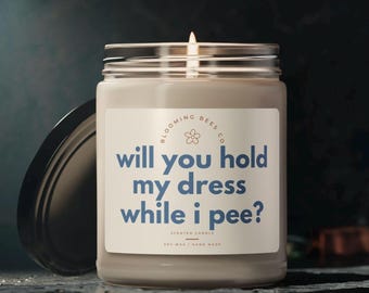 Peut inclure: Une bougie parfumée en cire de soja dans un bocal en verre avec un couvercle noir. L'étiquette indique "will you hold my dress while i pee ?" en texte bleu. La bougie est de Blooming Bees Co.