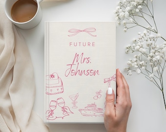 Op de afbeelding: Een lichtgekleurd boek met de woorden "FUTURE Mrs. Johnson" in roze script. De omslag bevat roze lijntekeningen van een handtas, champagneglazen, een cocktail, een jacht en een strik. Een kopje koffie en bloemen staan op de achtergrond.