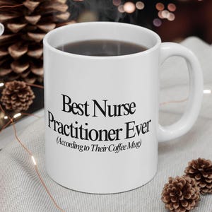 May include: White ceramic coffee mug with the text "Best Nurse Practitioner Ever (According to Their Coffee Mug)". The mug is filled with dark liquid and has a white handle. The background includes pinecones and string lights.